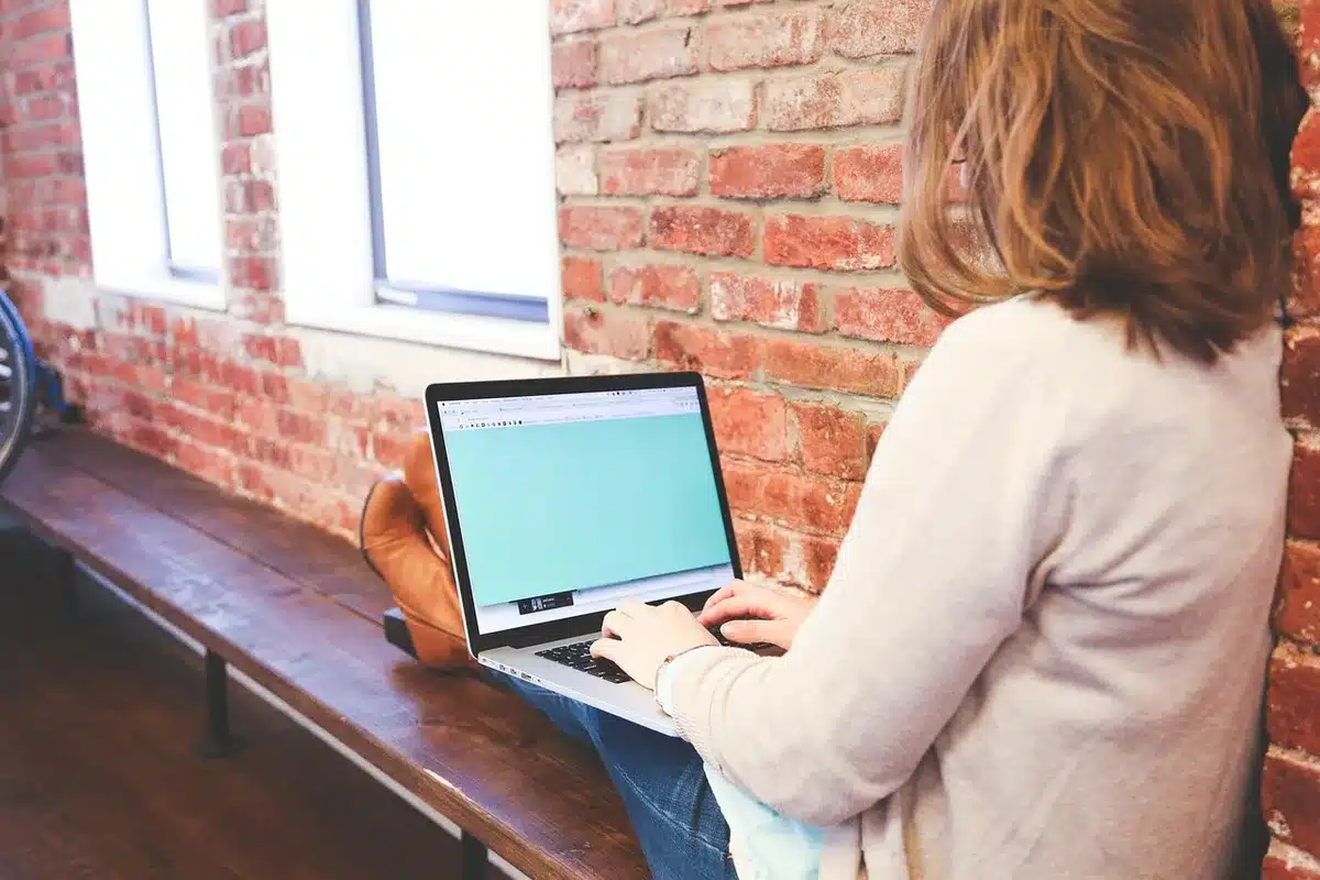 Hands typing on a laptop at a bright newsroom desk with a notepad and window view